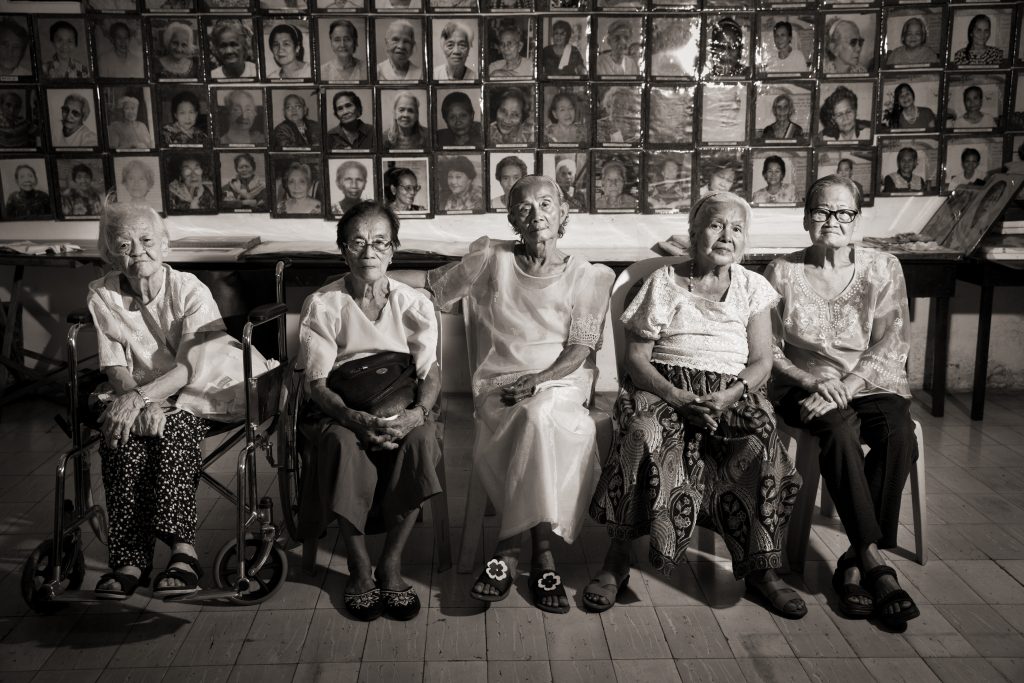 Four Filipino women, Lolas Remedios Tecson, Estela Adriatico, Narcisa Claveria, Felicidad delos Reyes and Estelita Dy, sit in chairs beside each other in front of a wall covered in photographs of the faces of other survivors. Photo taken on April 28, 2019 by Cheryl Diaz Meyer.